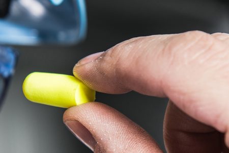 Worker inserting yellow foam earplug while wearing safety glasses, representing Raxwell ear protection products